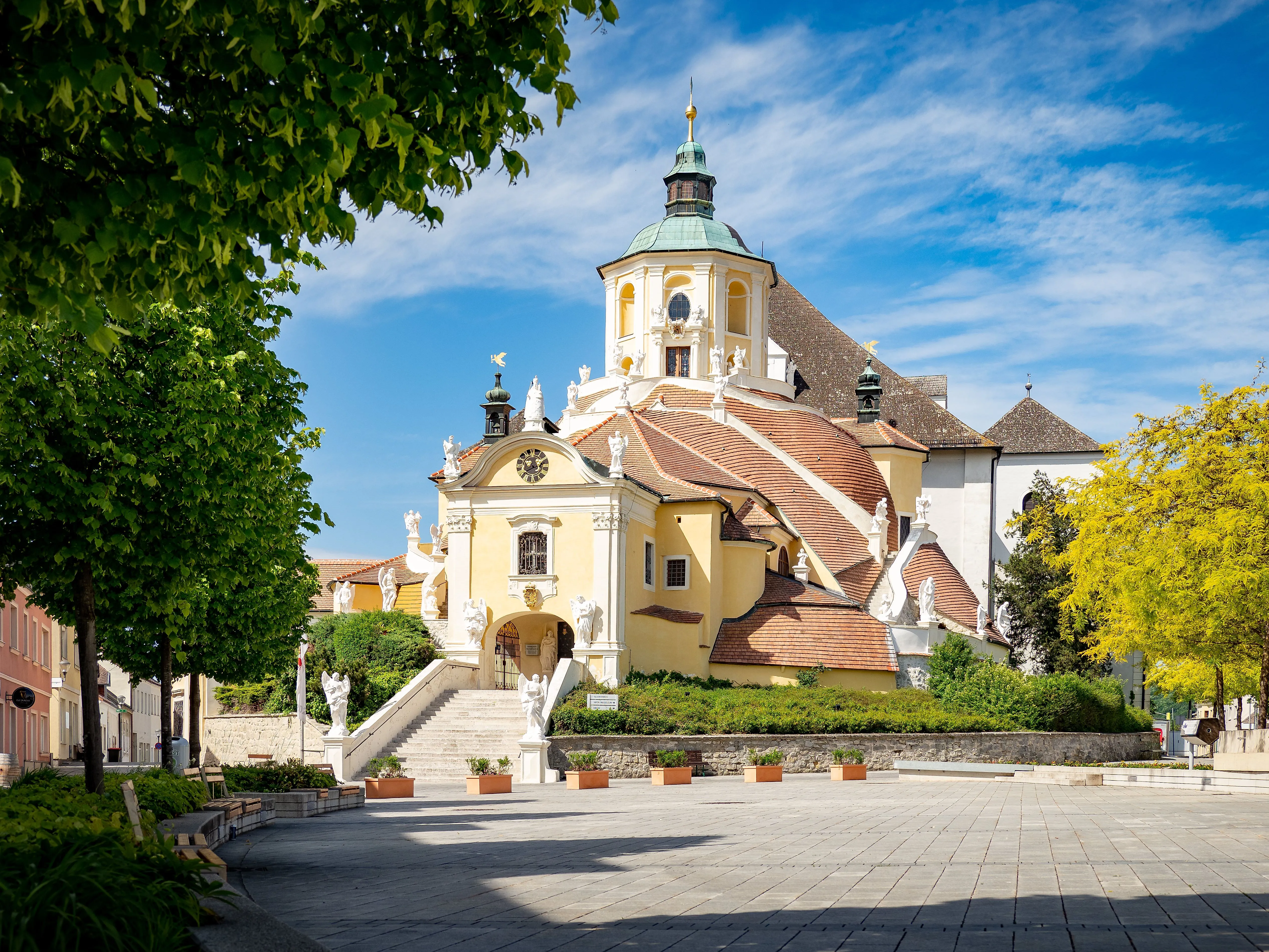 Die barocke Bergkirche in Eisenstadt mit ihren charakteristischen Türmen und Statuen, umgeben von blühendem Grün und blauem Sommerhimmel.
