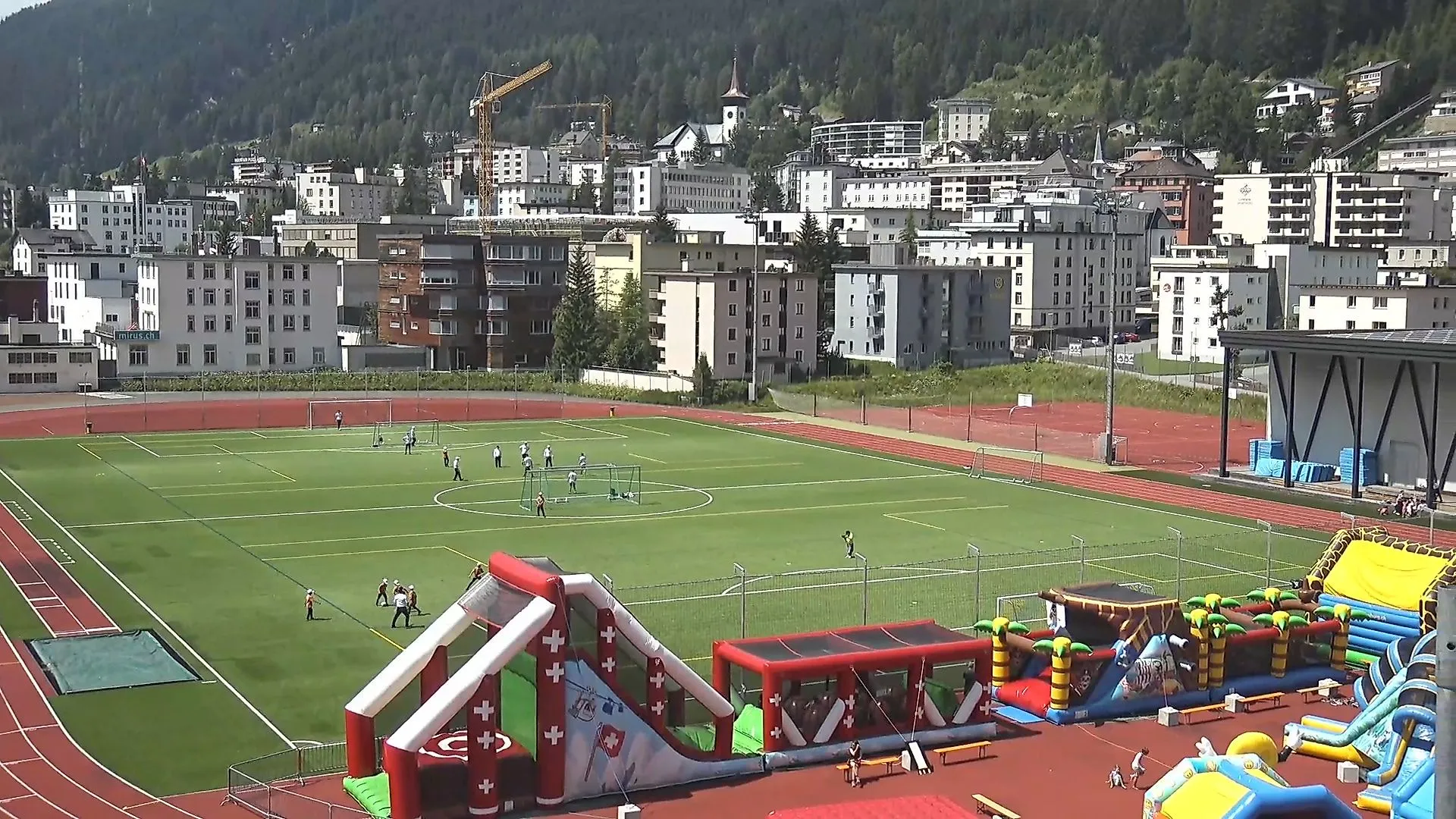 Sportzentrum Davos-Platz im Sommer mit Blick auf Kunstrasenplatz, auf dem Kinder Fußball spielen, und bunte Hüpfburgen im Vordergrund, umgeben von den Gebäuden und Bergen der Stadt.