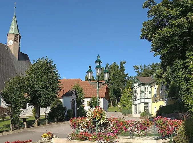 Letné námestie v Mönichkirchene s kvetinovými záhonmi, historickými budovami a modrou oblohou.