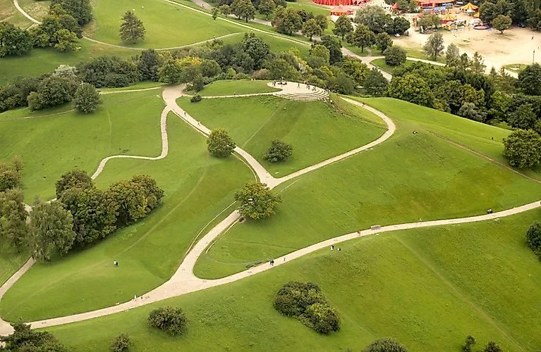 Luftaufnahme des Englischen Gartens in München mit geschwungenen Wegen, sanften Hügeln und verstreuten Bäumen an einem sonnigen Tag.