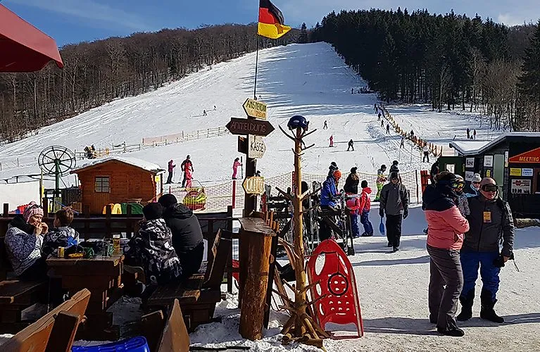 Día invernal en la estación de esquí familiar de Schlossberg, en Medebach, con esquiadores, trineístas y visitantes delante de la cabaña. La pista de esquí y un telesilla al fondo.