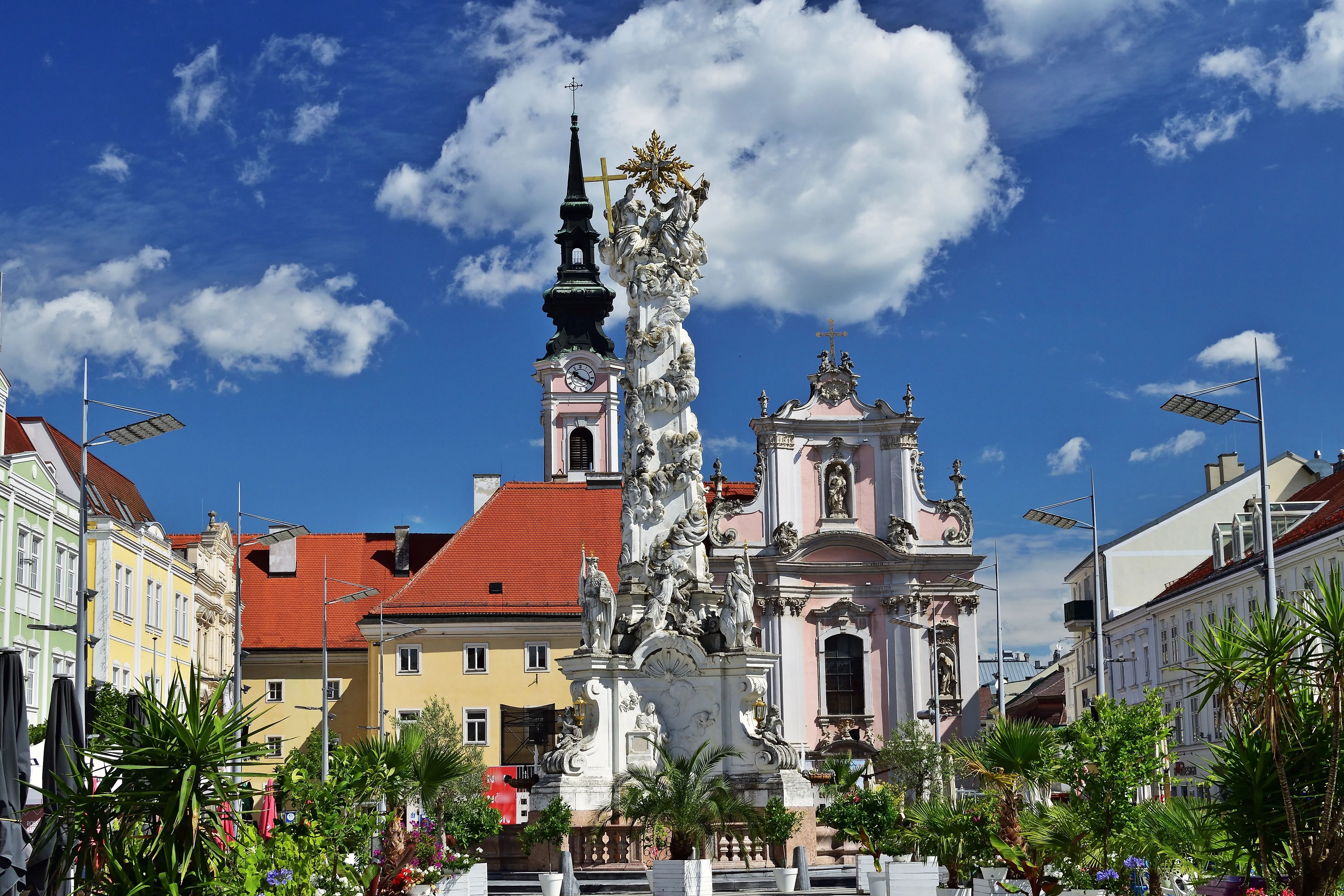 Historische Dreifaltigkeitssäule vor der barocken Franziskanerkirche im Zentrum von St. Pölten, umgeben von farbenfrohen Bürgerhäusern und sommerlichem Blumenschmuck.