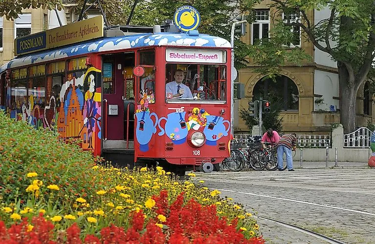 Bunte Straßenbahn des Ebbelwei-Expresses in Frankfurt mit fröhlicher Bemalung, im Vordergrund blühende Sommerblumen.