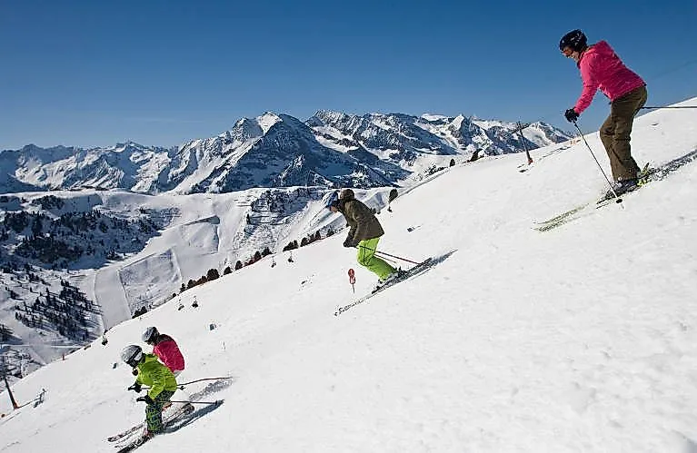 Familie fährt gemeinsam Ski auf einer sonnigen Piste mit herrlichem Alpenpanorama im Hintergrund.