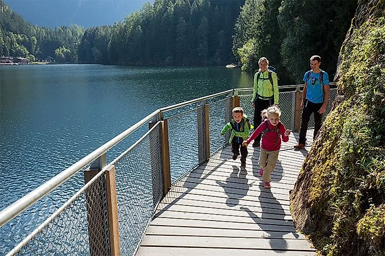 Paseos familiares por una pasarela de madera a lo largo del lago Piburger See, en el Tirol, con vistas al agua y el bosque de fondo.