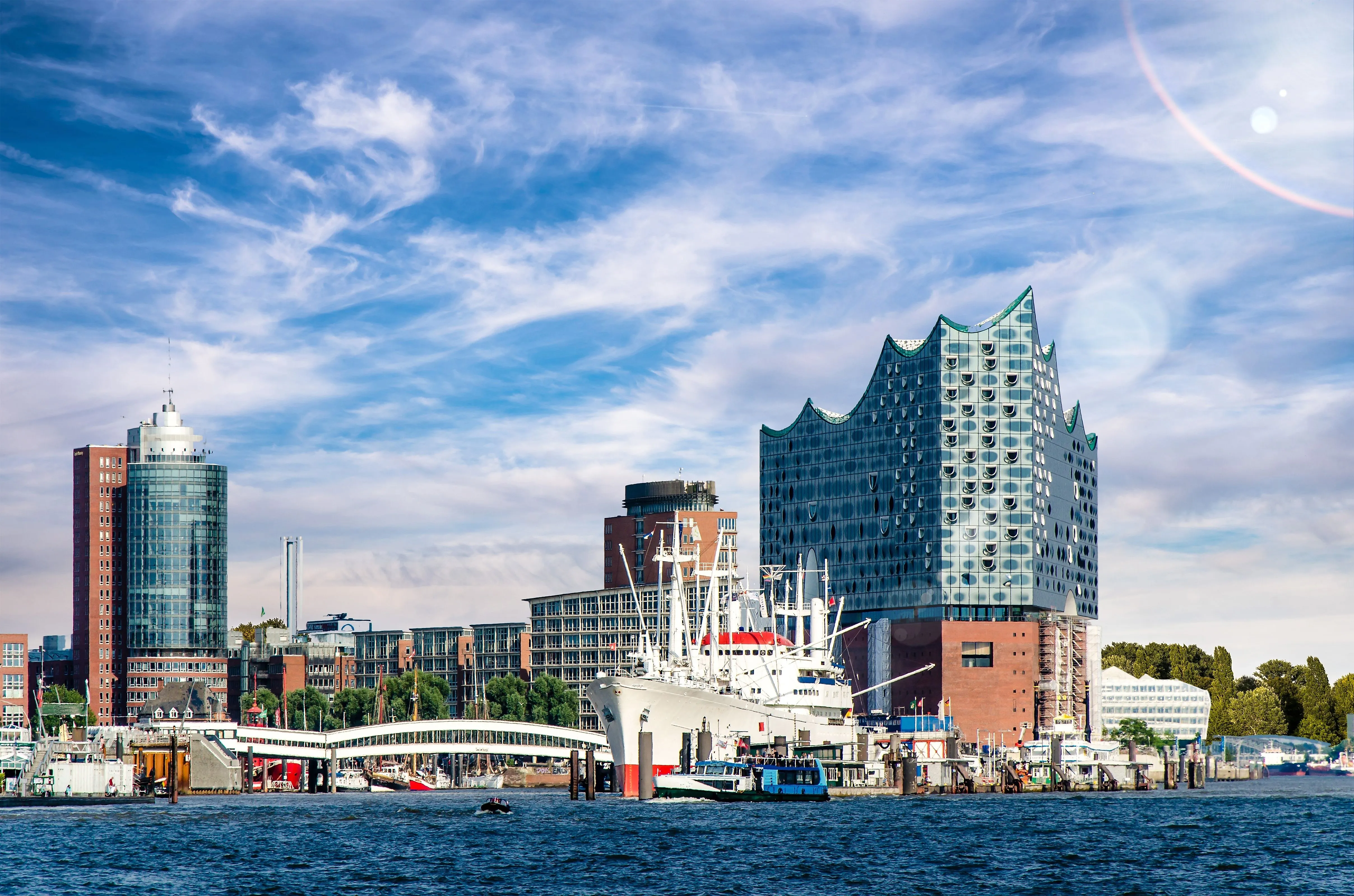 Panoramaaufnahme des Hamburger Hafens mit der modernen Elbphilharmonie und dem Museumsschiff Cap San Diego vor blauem Himmel.
