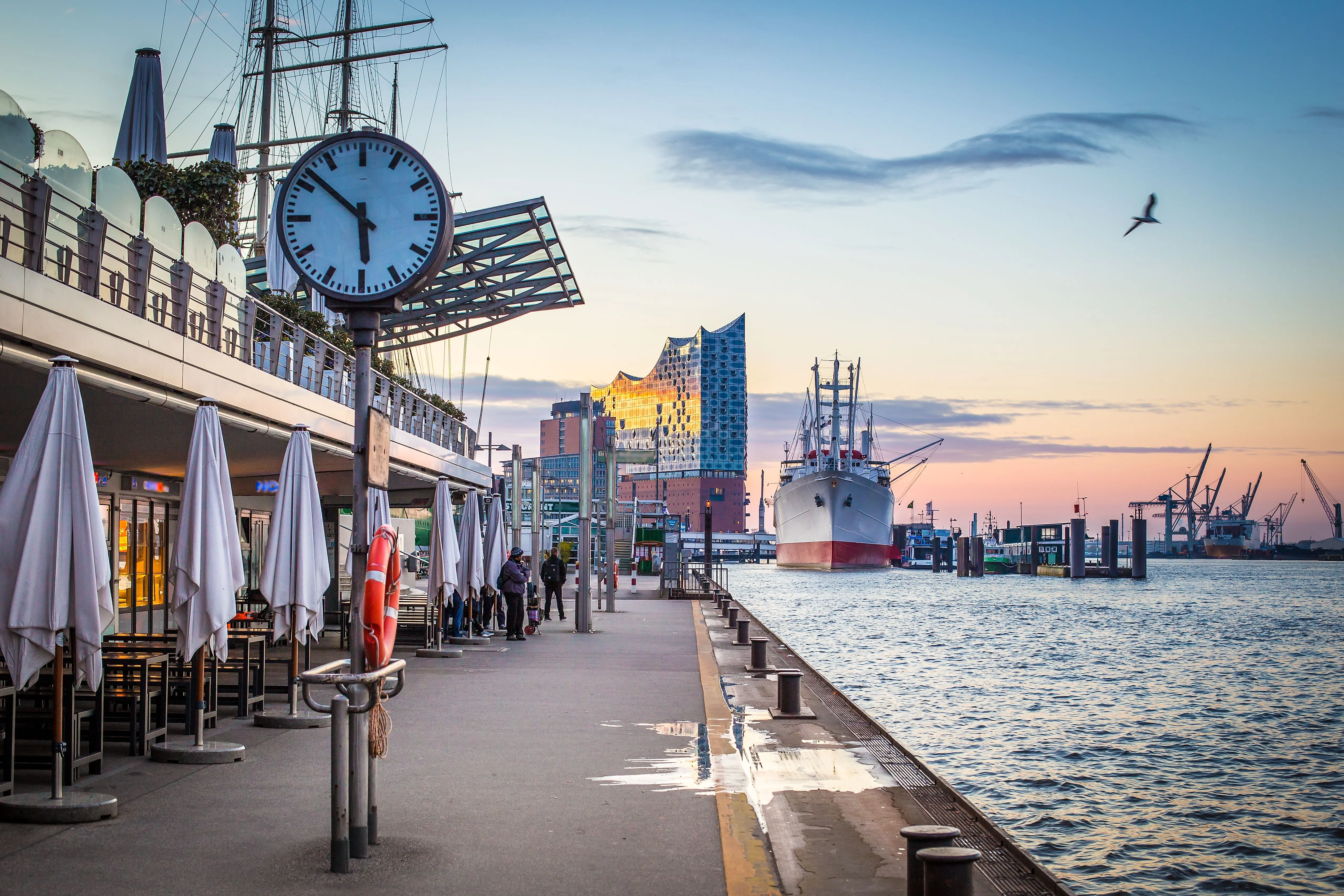 Spazierweg entlang der Hamburger Landungsbrücken mit Blick auf das Museumsschiff Cap San Diego und die beleuchtete Elbphilharmonie bei Sonnenuntergang.
