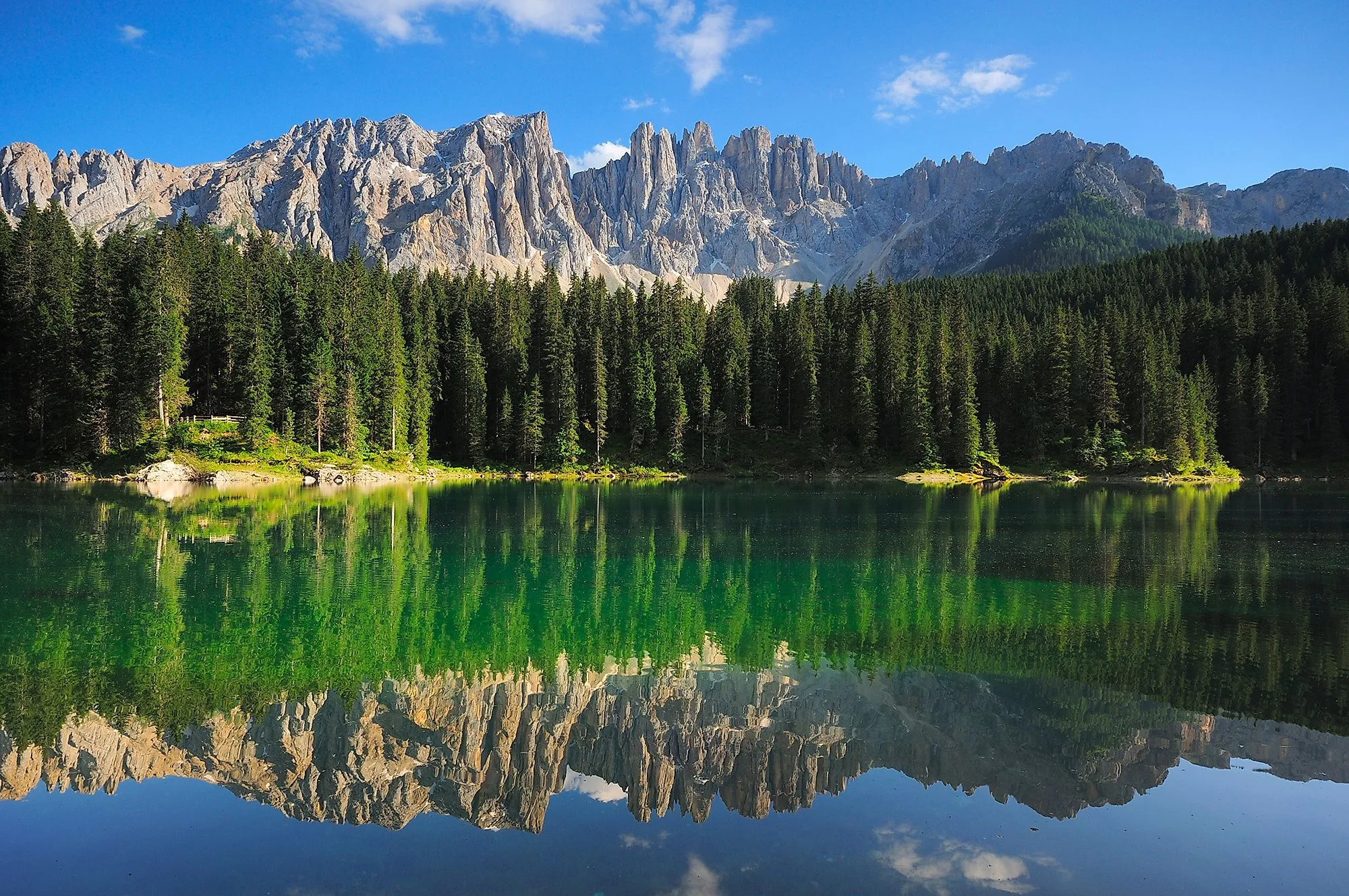 Kristallklarer Karersee mit Spiegelung des Latemar-Gebirges in Südtirol, umgeben von dichtem Wald und blauen Himmel.