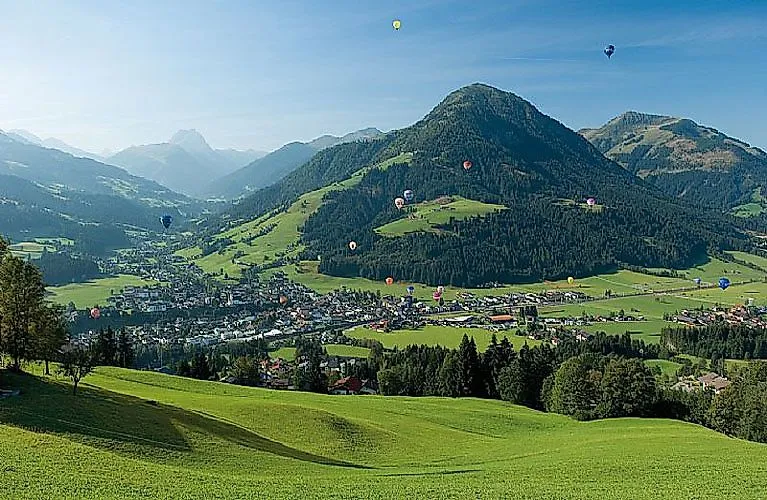 Sommerpanorama von Kirchberg in Tirol mit grünen Almwiesen, umliegenden Bergen und zahlreichen Heißluftballons am Himmel.