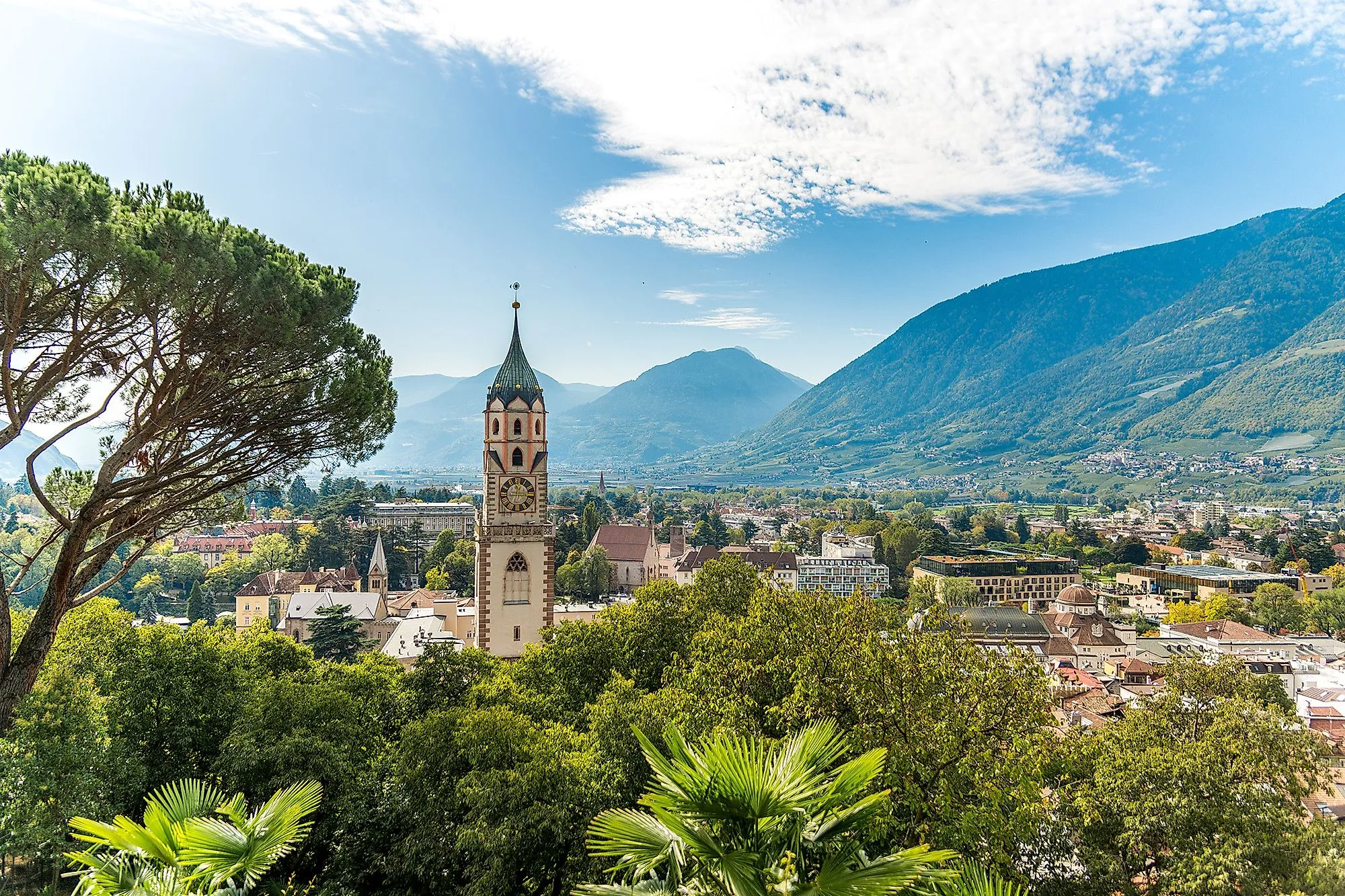 Panoramablick auf die Stadt Meran in Südtirol mit Kirchturm und umliegenden Bergen an einem sonnigen Tag