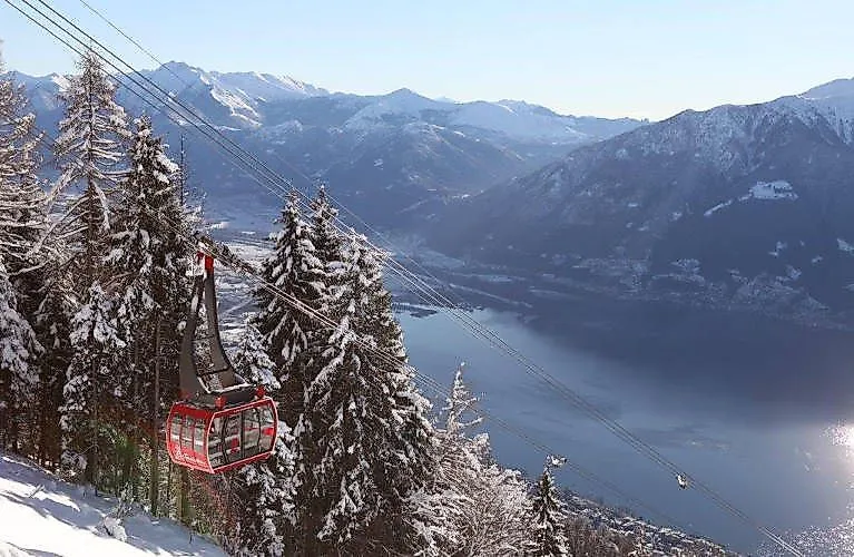 Rote Luftseilbahn fährt im Winter durch verschneiten Nadelwald mit Blick auf den Lago Maggiore und die umliegenden Alpen bei Minusio.