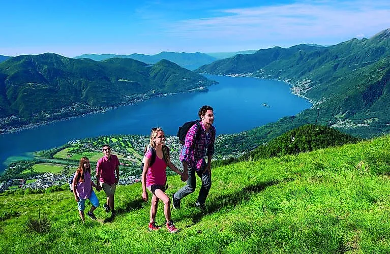 Wandergruppe mit Familie genießt sonnigen Sommertag auf grüner Almwiese mit Panoramablick auf den Lago Maggiore bei Minusio.