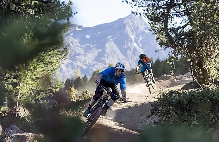 Zwei Mountainbiker fahren auf einem Trail durch den Wald in Längenfeld mit Bergkulisse im Hintergrund.