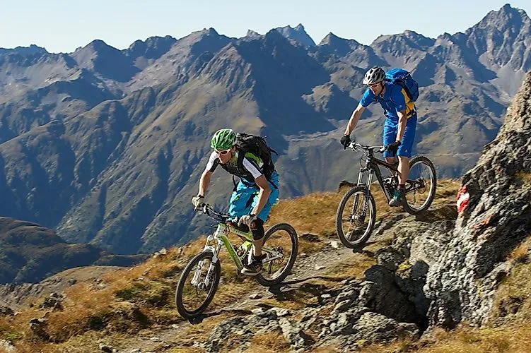 Two mountain bikers ride on an alpine trail above Ischgl, surrounded by imposing mountain ridges in the Tyrolean alpine landscape.