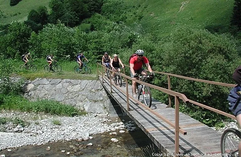 Groupe de VTTistes traversant un étroit pont en bois au-dessus d'un ruisseau de montagne à Berwang, entouré d'un paysage alpin verdoyant en été.