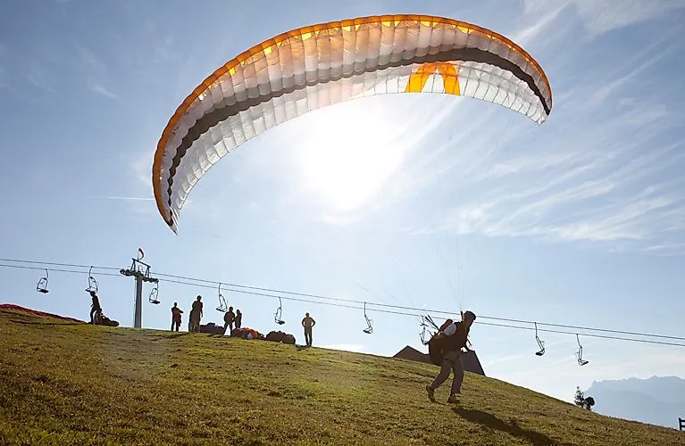 Gleitschirmflieger startet auf grüner Wiese bei Kössen mit Blick auf Seilbahn und Berge an einem sonnigen Sommertag