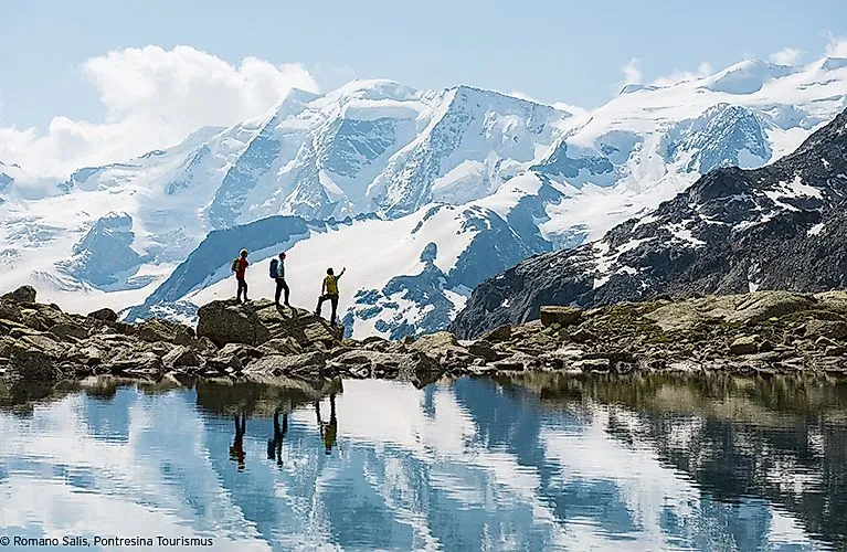 Drei Wanderer stehen auf Felsen am See in der Region Pontresina, umgeben von verschneiten Bergen und Spiegelung im Wasser.