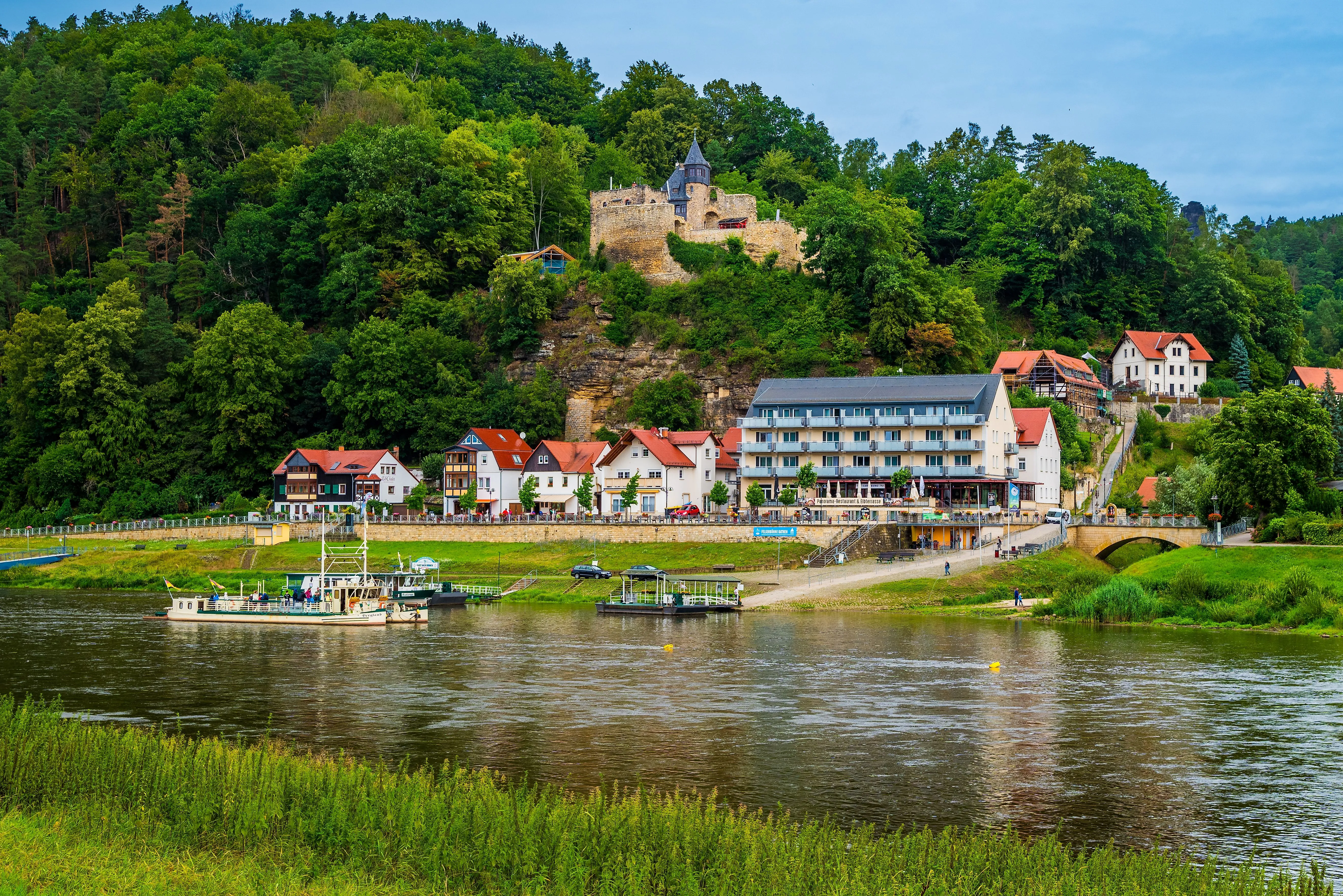 Blick auf das idyllische Elbufer mit der Fähre in Rathen, umgeben von Fachwerkhäusern und der malerischen Kulisse der Burg Altrathen.