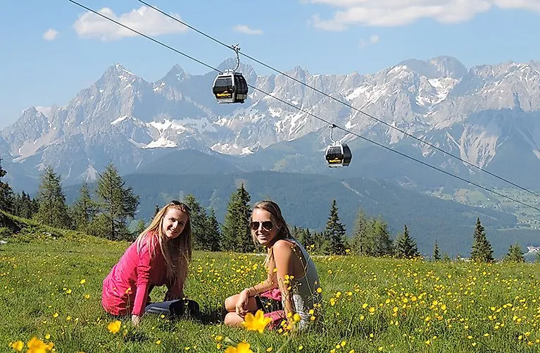 ALT-Text:Zwei junge Frauen genießen im Sommer die Aussicht auf der Reiteralm, mit Blick auf die imposanten Dachstein-Berge und vorbei fahrenden Gondeln.