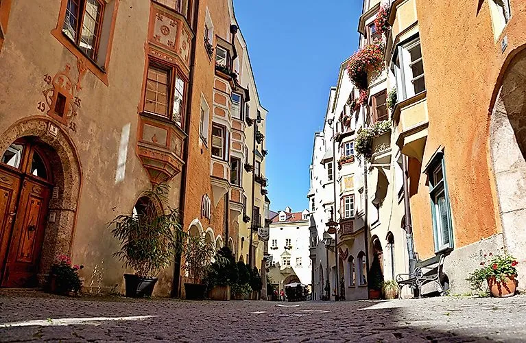 Enge romantische Gasse in Hall in Tirol mit farbenfrohen historischen Fassaden, Erkern, Blumenschmuck und Kopfsteinpflaster bei strahlendem Sonnenschein.