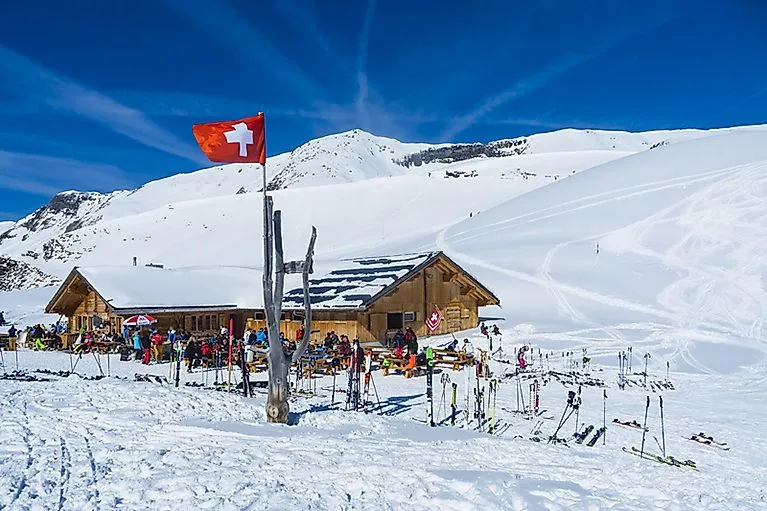 Skihütte mit Schweizer Flagge auf einer verschneiten Skipiste bei Grindelwald, umgeben von Wintersportlern unter strahlend blauem Himmel.