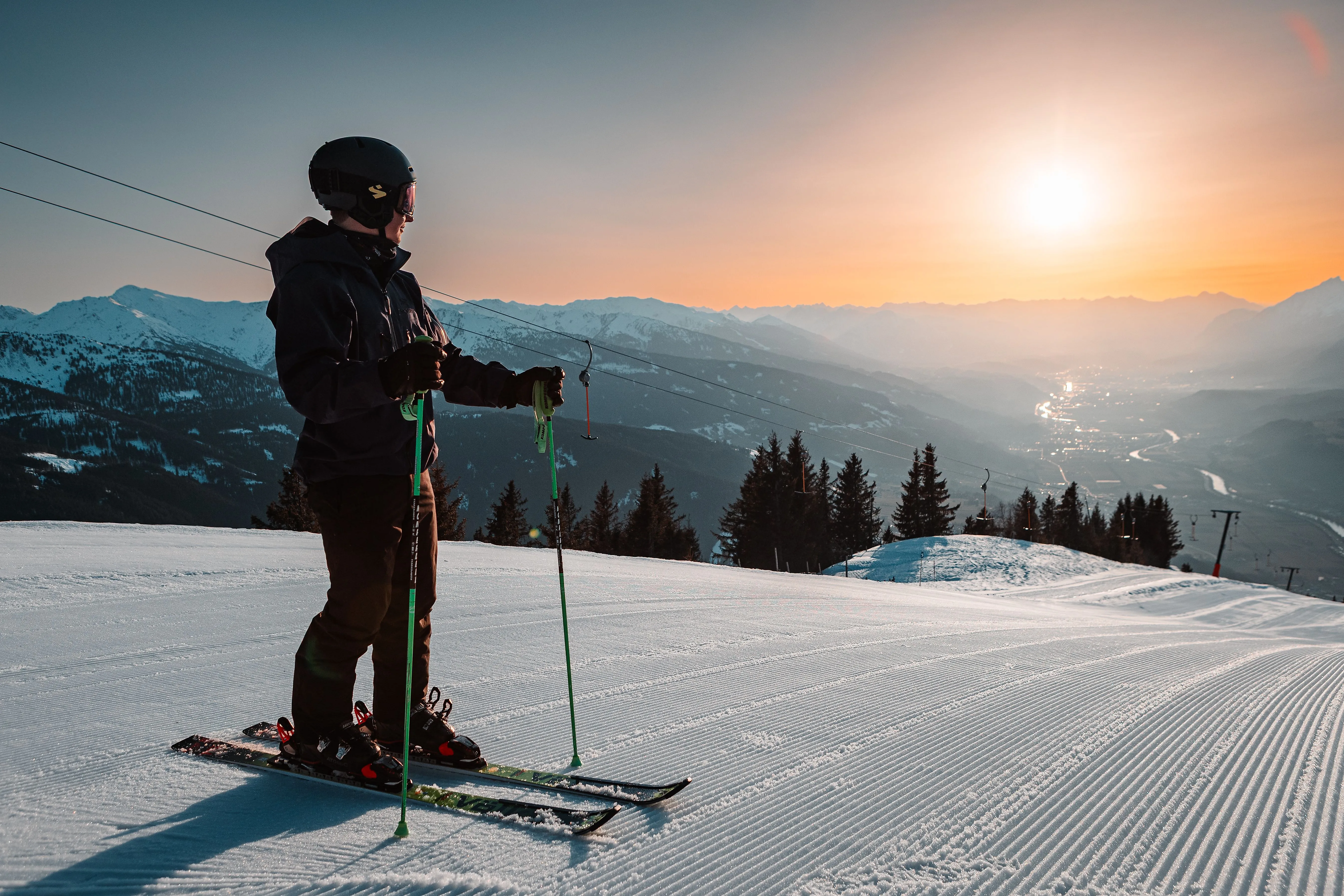 Skifahrer auf präparierter Piste bei Sonnenuntergang mit Blick auf verschneite Alpen und Tal im Hintergrund