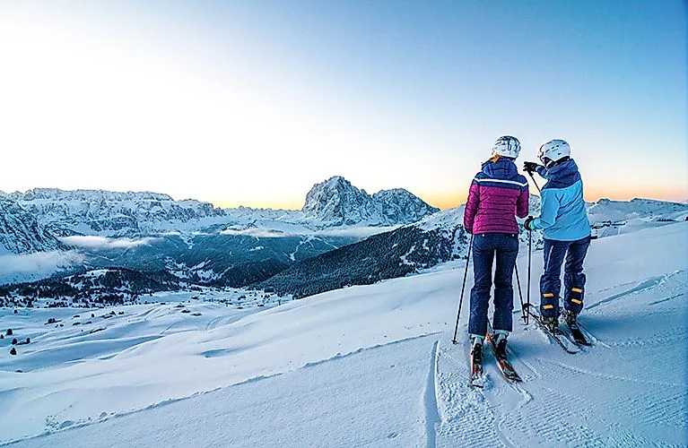 Twee skiërs op een besneeuwde piste met uitzicht op de Dolomieten bij zonsopgang