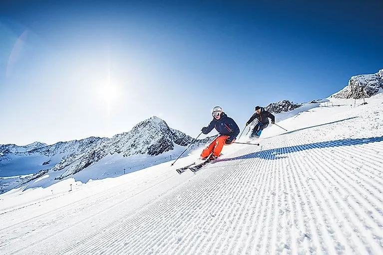 Zwei Skifahrer auf einer frisch präparierten Piste am Stubaier Gletscher mit strahlendem Sonnenschein und verschneiten Berggipfeln.