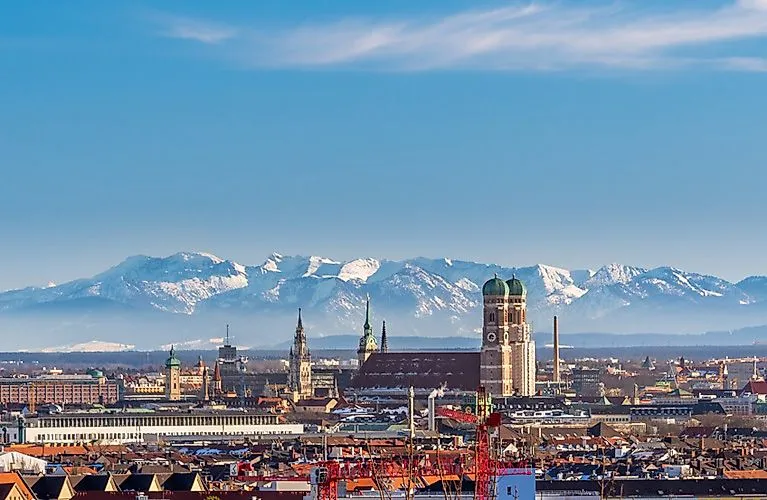 Blick auf die Skyline von München mit der Frauenkirche im Vordergrund und den schneebedeckten Alpen im Hintergrund bei klarem Himmel.