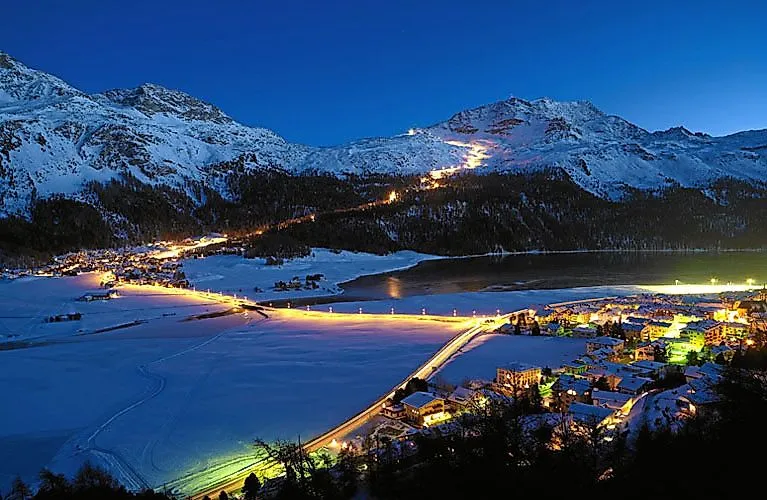 Winterliche Nachtaufnahme von St. Moritz mit beleuchteter Brücke und Skipisten, eingerahmt von schneebedeckten Alpen.