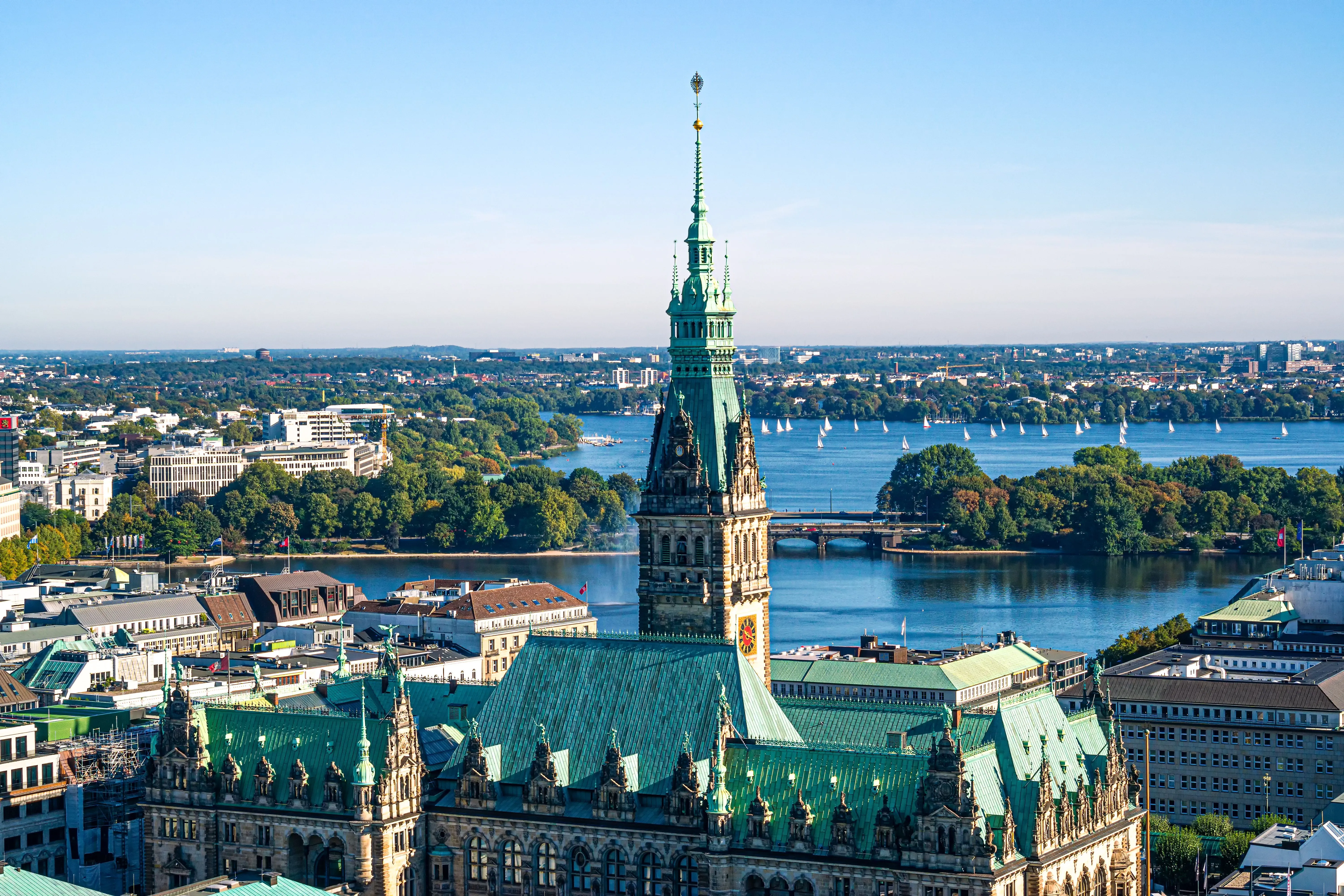 Blick über Hamburg mit dem historischen Rathaus im Vordergrund und der Außenalster im Hintergrund, auf der zahlreiche Segelboote unterwegs sind.