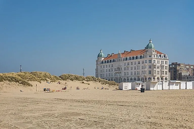 Helles Sandstrandpanorama von Zeebrugge mit einigen Badegästen und einem historischen weißen Hotelgebäude mit grünen Kuppeldächern im Hintergrund bei blauem Himmel.
