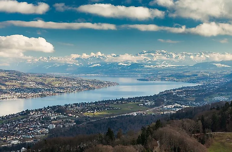 Weitblick über den Zürichsee mit angrenzender Stadtlandschaft und schneebedeckten Alpen im Hintergrund.
