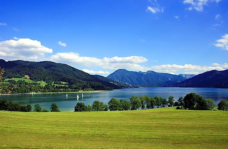 Weite Sommerlandschaft mit Blick auf den Tegernsee, gesäumt von grünen Wiesen und bewaldeten Bergen.