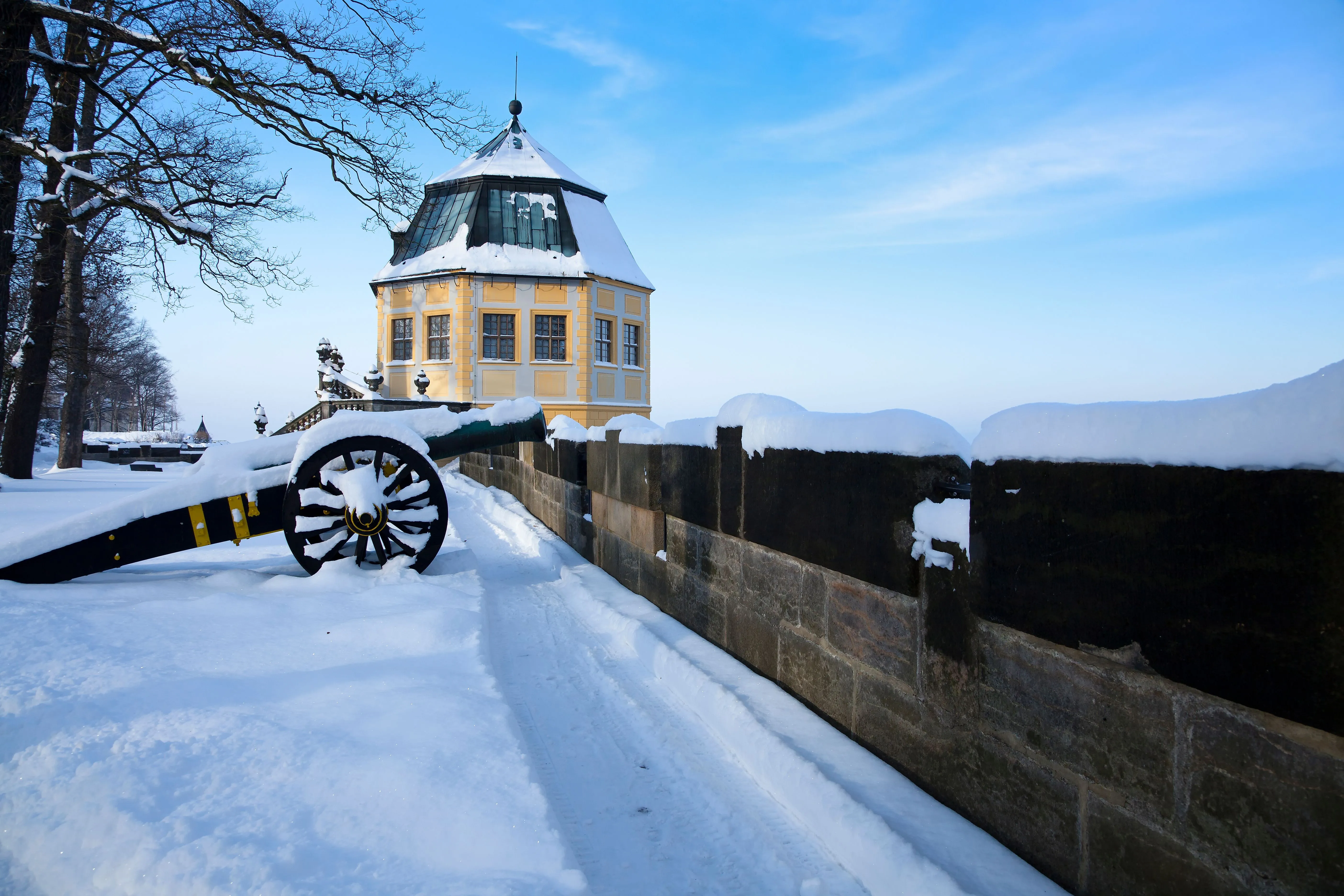 Historische Kanone und schneebedeckte Gebäude der Festung Königstein bei Bad Schandau mit Blick auf das Elbsandsteingebirge im Winter.