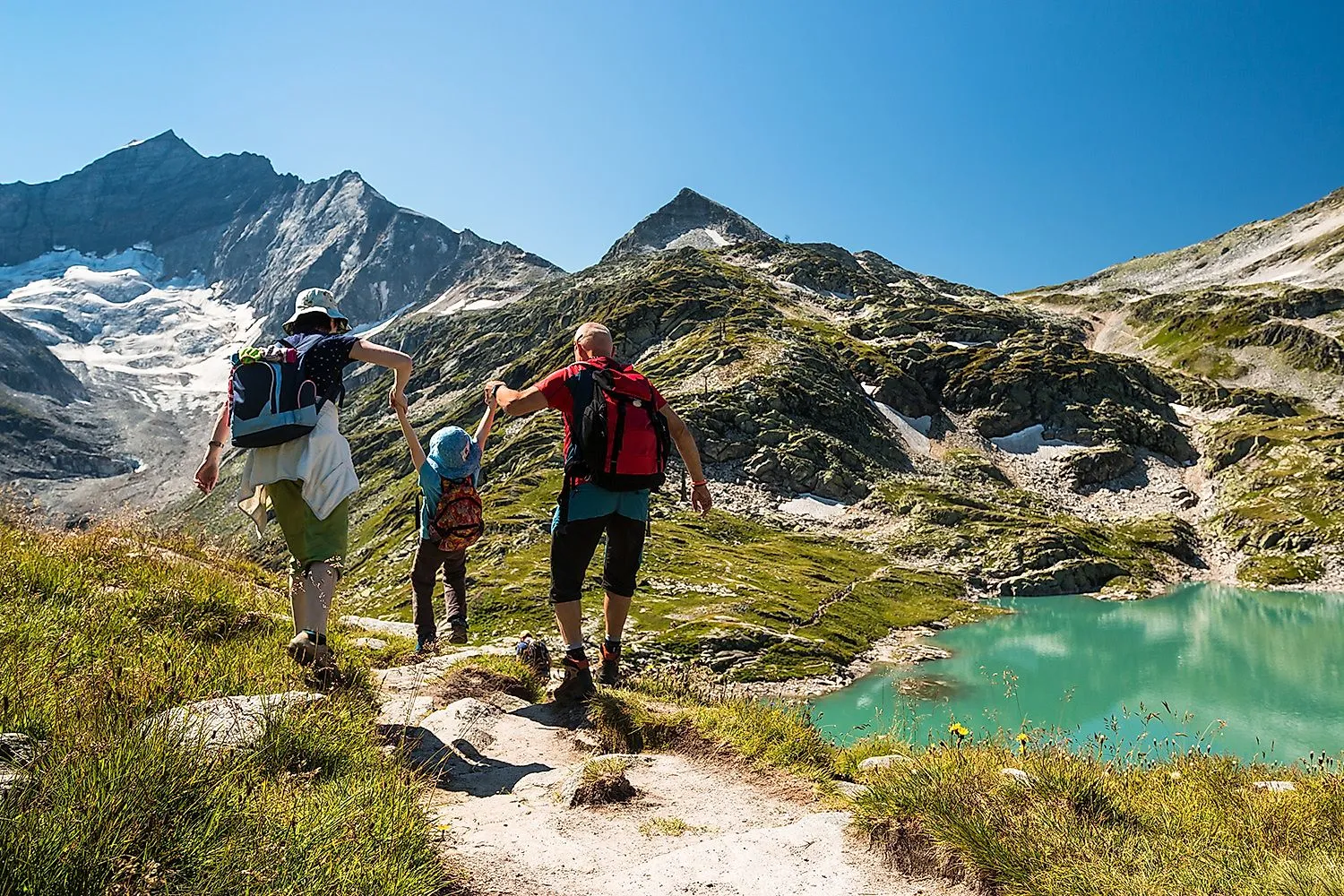 Familie mit Kind auf einer Bergwanderung entlang eines alpinen Wanderwegs in den österreichischen Alpen, mit Gletscher, Bergsee und sonnigem Wetter im Hintergrund.