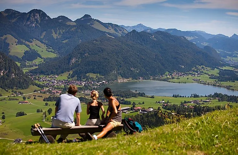 Wanderer auf einer Bank mit Blick auf das Tal, den Walchsee und die Tiroler Bergwelt bei schönem Wetter in Kössen