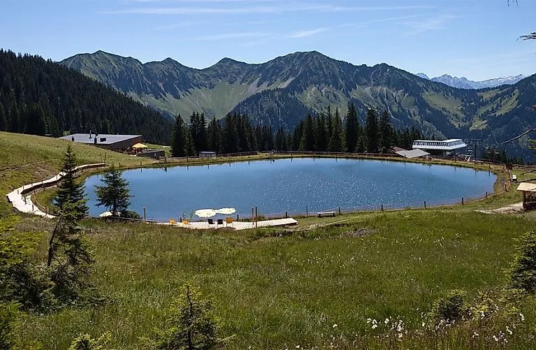 Almlandschaft in Laterns mit idyllischem Bergsee, umgeben von grünen Wiesen und bewaldeten Bergen im Sommer.