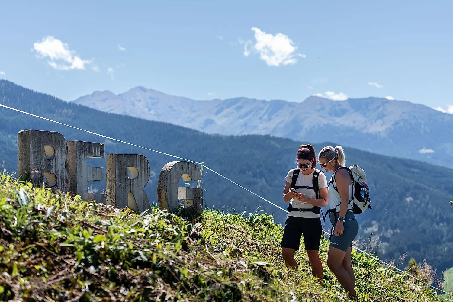 Zwei Wanderinnen am Berg mit Blick auf Alpen und Schriftzug Berg im Vordergrund bei sonnigem Wetter