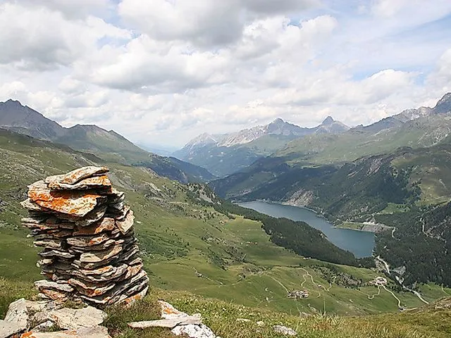 Panoramablick auf die grüne Berglandschaft von Savognin mit einem Steinmännchen im Vordergrund und einem tiefblauen Bergsee in der Ferne.