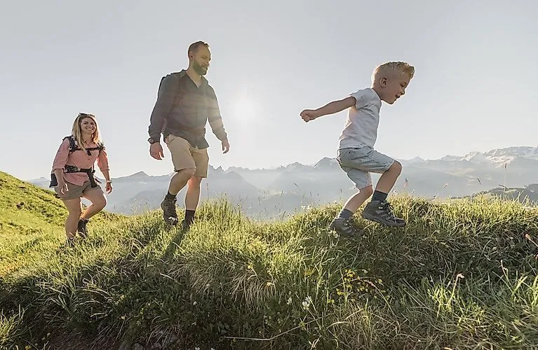 Familie beim Wandern auf sonnigen Bergwiesen oberhalb von Saanenmöser mit herrlichem Ausblick auf die Alpen.