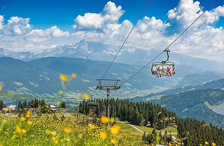 Sommerliche Berglandschaft in Flachau mit blühenden Wiesen, Seilbahn und Blick auf die Alpen im Hintergrund.