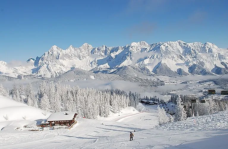 Verschneite Winterlandschaft auf der Reiteralm mit Skifahrern auf der Piste und Blick auf das Dachsteinmassiv im Hintergrund.
