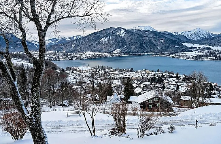 Winterliche Panoramaaufnahme des Tegernsees mit schneebedeckten Häusern und Bergen im Hintergrund.