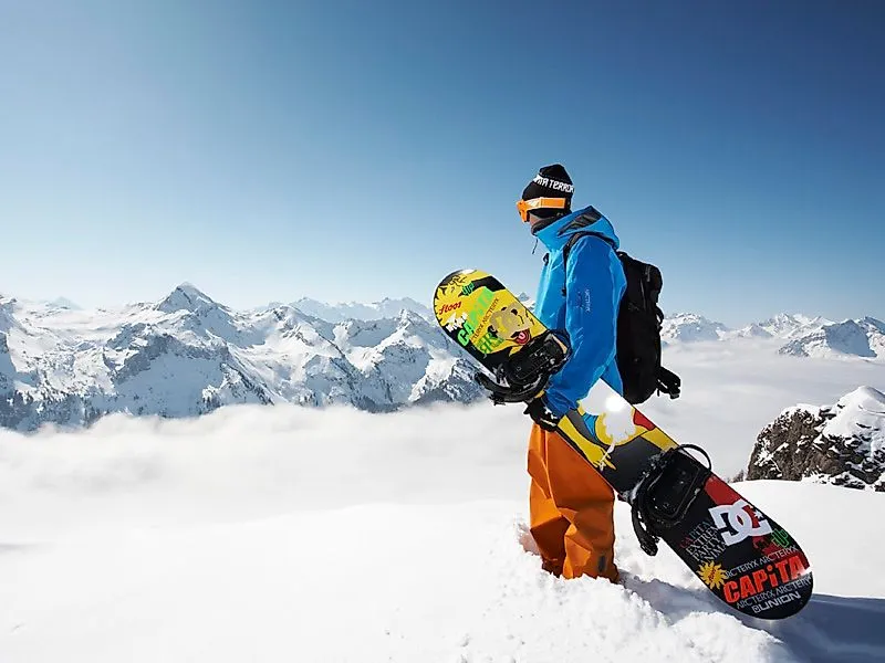 Snowboarder mit bunter Ausrüstung steht im Pulverschnee vor alpinem Panorama bei Stoos
