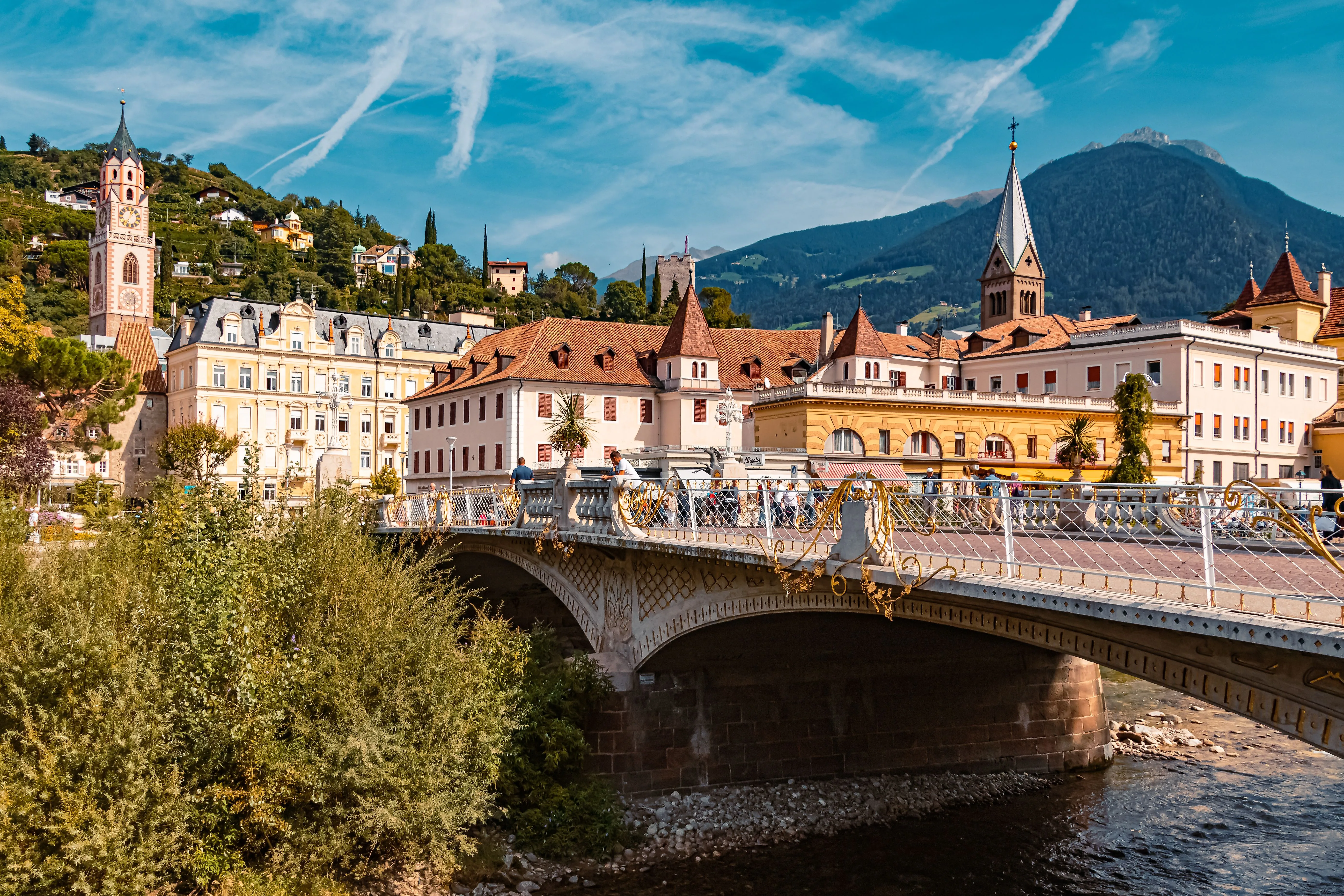 Blick auf die historische Altstadt von Meran mit der Postbrücke und den Bergen im Hintergrund an einem sonnigen Sommertag.