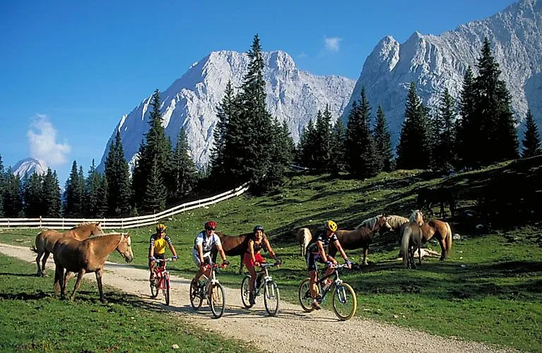 Štyria cyklisti prechádzajú cez alpskú lúku neďaleko Ehrwaldu, obklopenú koňmi a impozantnými horami regiónu Zugspitze.