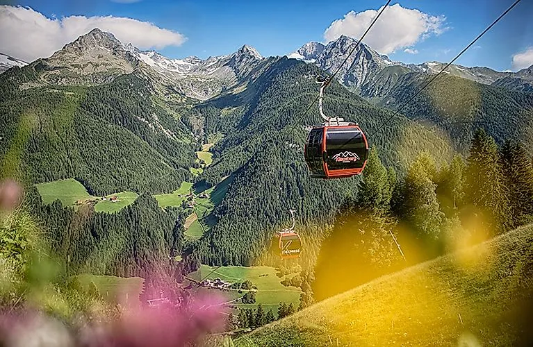Rote Gondeln schweben über eine grüne Sommerlandschaft mit blühenden Wiesen und hohen Bergen im Ahrntal am Klausberg.