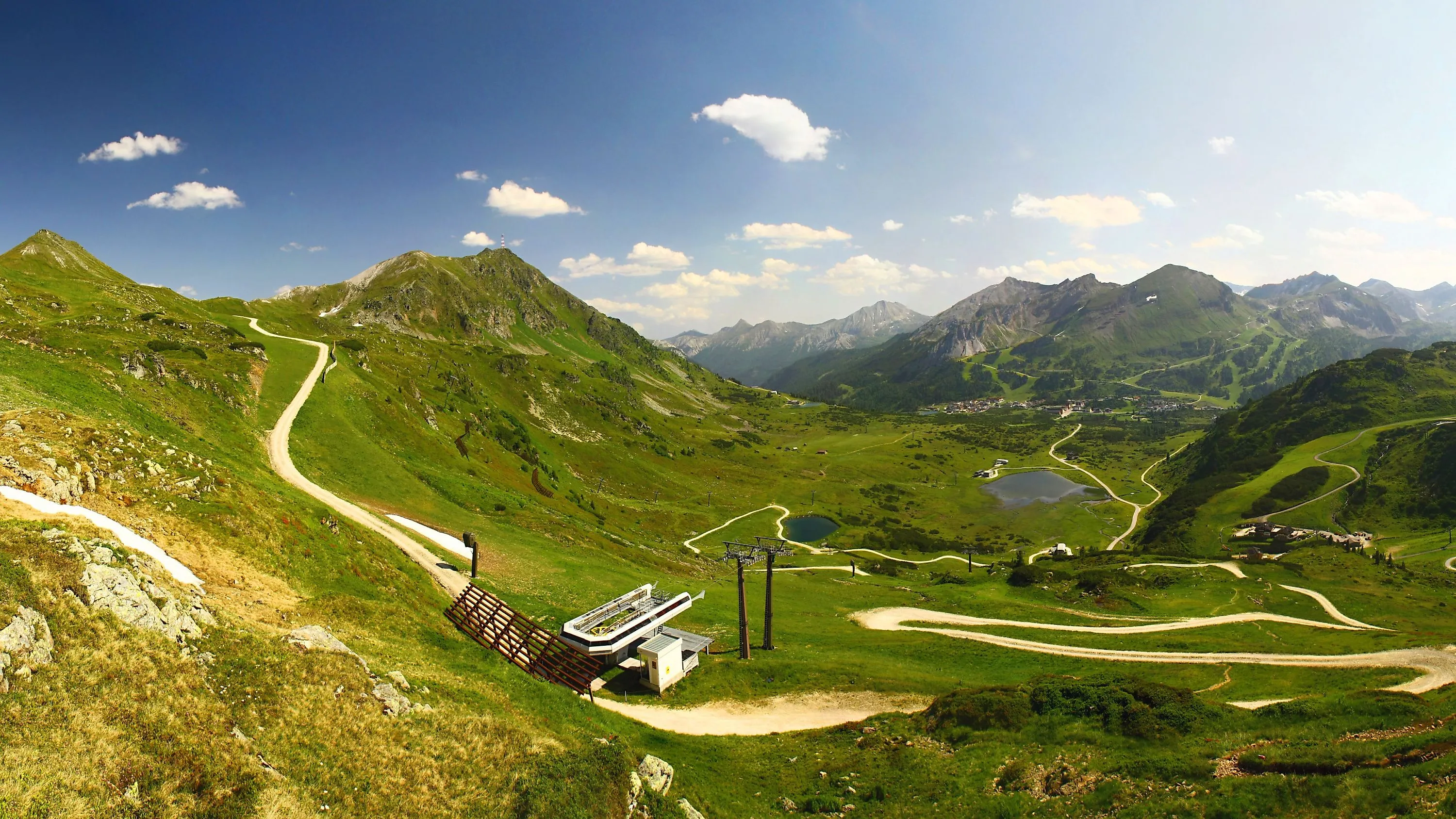 Sommerliche Panoramaaufnahme von Obertauern mit sanften Almwiesen, kurvigen Wanderwegen und einem klaren Bergsee, eingebettet in die imposante Bergwelt der Radstädter Tauern.