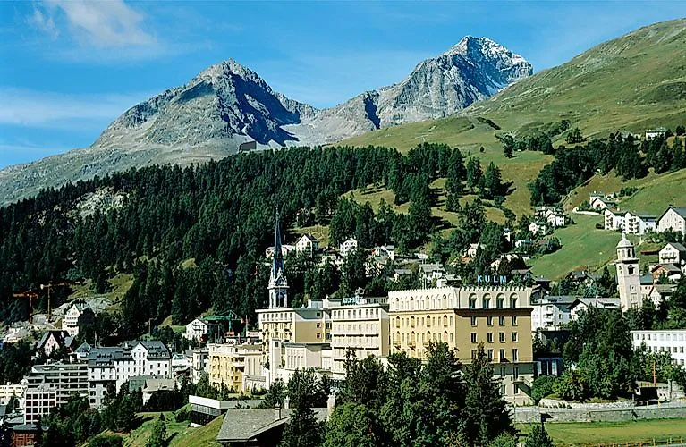 Zentrale Ansicht des Ortes St. Moritz mit dem historischen Kulm Hotel und den umliegenden Bergen an einem sonnigen Sommertag.